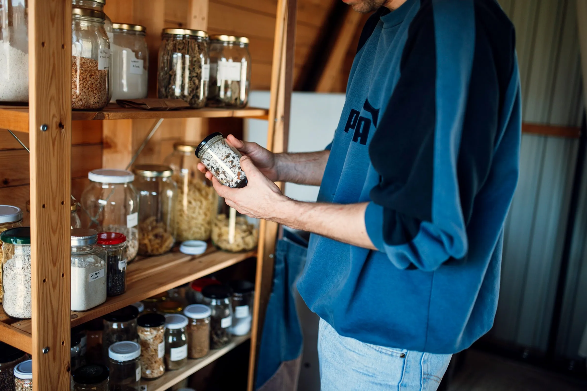 A person lifting a glass jar from a wooden kitchen shelf stacked with storage jars