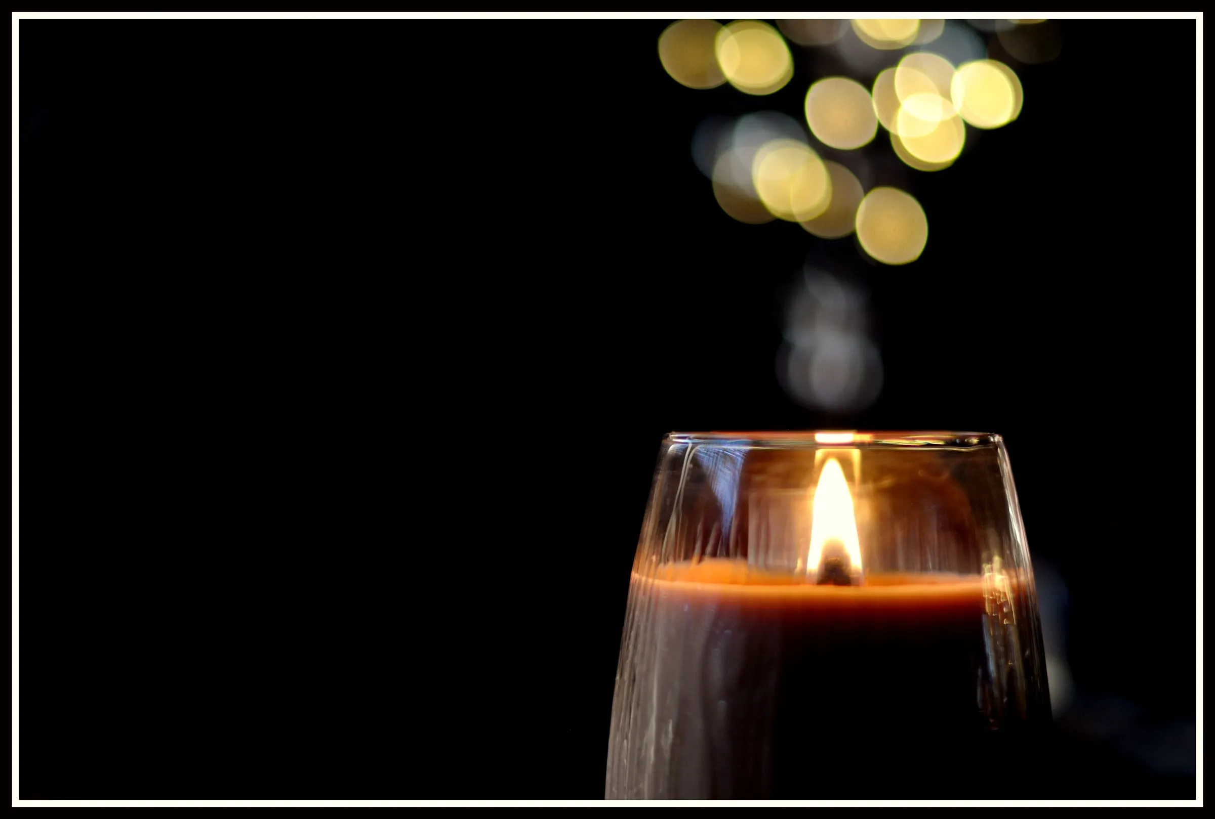 A single candle burning in a glass on a dark table, with warm lights out of focus behind it