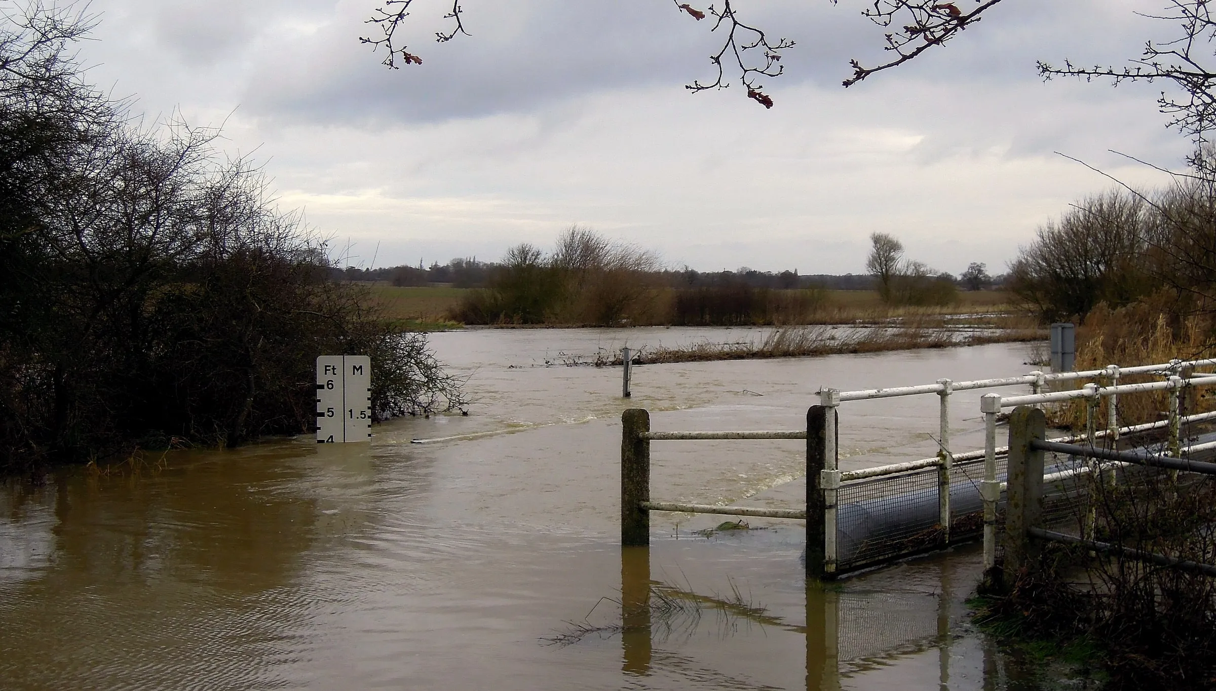 A swollen brown river in flood at Buttsbury Ford in Essex, water up over the field gate and depth marker